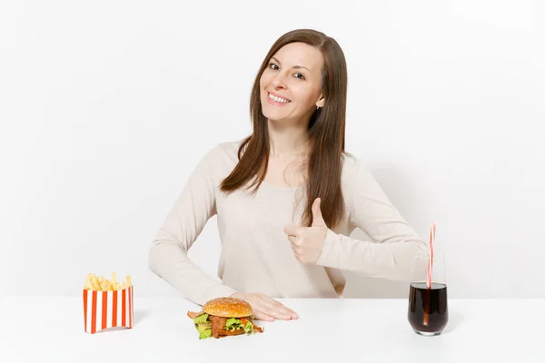 Beautiful woman shows thumbs up at table with burger, french fries, cola in glass bottle isolated on white background. Proper nutrition or American classic fast food. Advertising area with copy space. — Stock Photo, Image