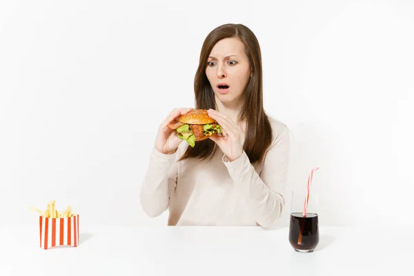 Jeune femme insatisfaite assise à table avec hamburger, frites, cola en bouteille de verre isolé sur fond blanc. Une bonne nutrition ou de la restauration rapide classique américaine. Espace publicitaire avec espace de copie — Photo