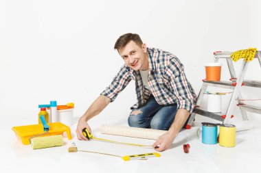Rulo duvar kağıdı, ölçü birimi teyp, kalem, yenileme daire oda için aygıtlar ile katta oturan genç adam beyaz arka plan üzerinde izole. Aksesuarlar, boyama araçlarını yapıştırma. Onarım ev concept