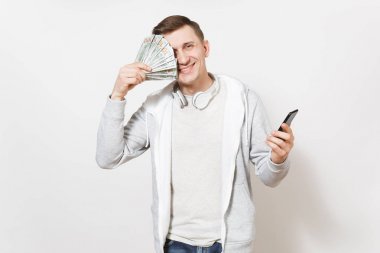 Genç adam t-shirt, boynuna kulaklıklarla ışık sweatshirt içinde gülümseyen yüz dolar, nakit para bohça ile kapak ve beyaz arka plan üzerinde izole elinde cep telefonu tut. Başarı kavramı