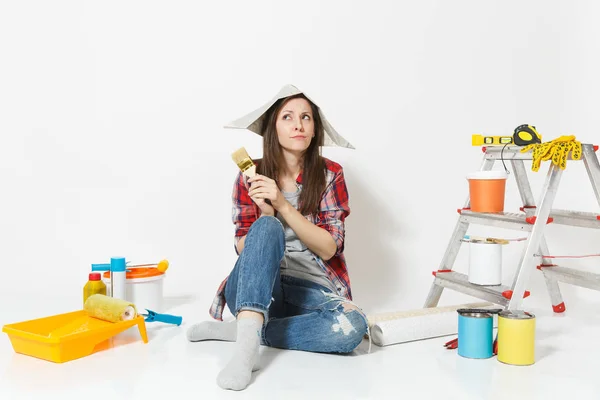 Mulher bonito em chapéu de jornal sentado no chão com escova, instrumentos para renovação apartamento quarto isolado no fundo branco. Papel de parede, acessórios para colar, ferramentas de pintura. Reparação home concept . — Fotografia de Stock