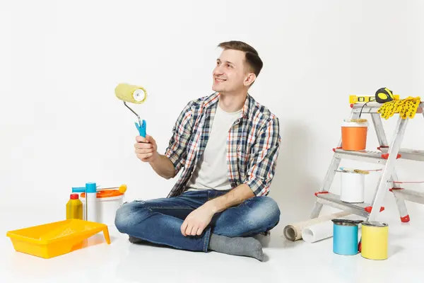 Homem de roupas casuais sentado no chão com rolo de tinta, instrumentos para renovação apartamento quarto isolado no fundo branco. Papel de parede, acessórios de colagem, ferramentas de pintura. Reparação home concept . — Fotografia de Stock