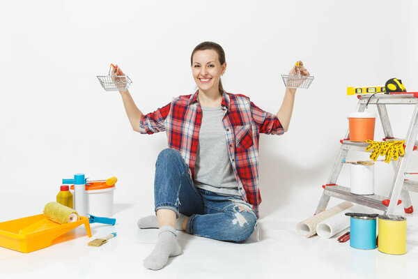 Woman holds metal grocery baskets for shopping, sits on floor with instruments for renovation apartment isolated on white background. Wallpaper, accessories, tools for painting. Repair home concept.