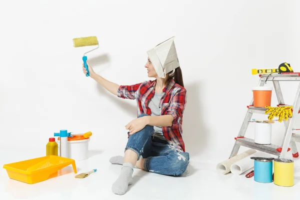 Linda mujer en sombrero de periódico sentado en el suelo con rodillo de pintura, instrumentos para apartamento de renovación aislado sobre fondo blanco. Accesorios de papel pintado para pegar, herramientas de pintura. Reparación concepto hogar — Foto de Stock