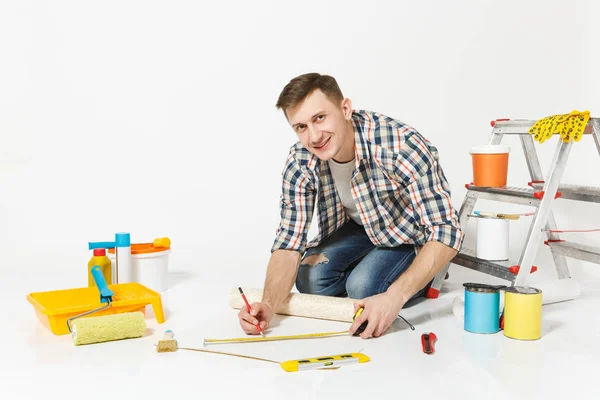 젊은 남자 롤 벽지, 측정 테이프, 연필, 재건축 아파트 룸을 위한 계기의 바닥에 앉아 흰색 배경에 고립. 액세서리, 도구 그림 붙이기 수리 홈 개념 — 스톡 사진
