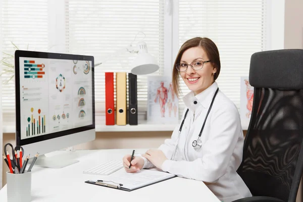 Young smiling woman sitting at desk, working on computer with medical documents in light office in hospital. Female doctor in medical gown, stethoscope in consulting room. Healthcare, medicine concept — Stock Photo, Image