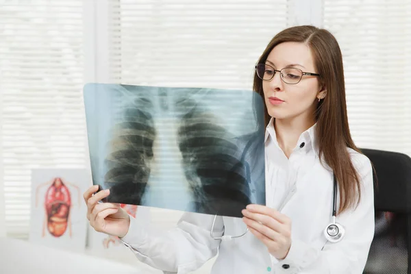 Ung självsäker kvinnliga läkare står vid skrivbord, röntgen av lungorna, fluorography, roentgen inom ljus kontor i sjukhuset. Kvinna i medicinsk klänning, stetoskop i samråd rum. Medicin-konceptet. — Stockfoto