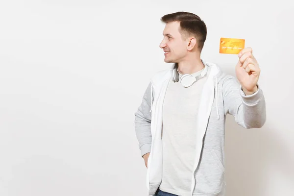 젊은 잘생긴 웃는 남자 학생 t-셔츠, 멀리에 고립 된 흰색 배경에 보이는 목 헤드폰으로 가벼운 운동복 카메라, 돈을 오렌지 신용 카드를 보여줍니다. 성공의 개념 — 스톡 사진
