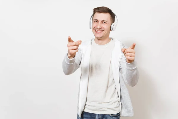 Joven guapo divertido hombre sonriente estudiante en camiseta y sudadera ligera escuchando música con auriculares inalámbricos blancos y muestra los dedos en la cámara aislados sobre fondo blanco. Concepto de emociones —  Fotos de Stock