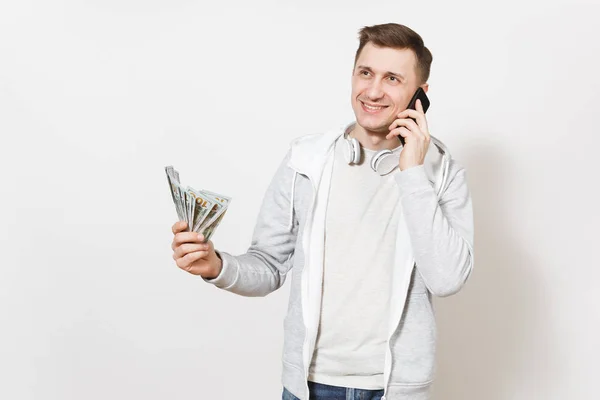 Молодой красивый улыбающийся мужчина в футболке, светлая толстовка с наушниками на шее, разговаривающий по мобильному телефону с пачкой долларов, наличные деньги в руке изолированы на белом фоне. Концепция успеха . — стоковое фото