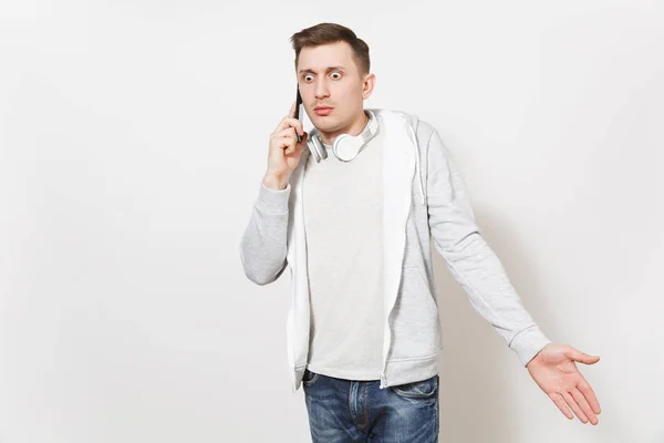 Mladý pohledný ustaraný muž student v tričku, lehká mikina s sluchátka kolem krku rozhovory na mobilním telefonu a vypadá zmatený ve studiu na bílém pozadí. Koncepce komunikace, emoce. — Stock fotografie