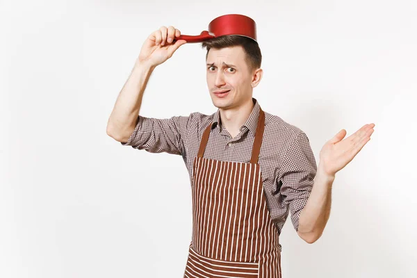 Young fun crazy sad man chef or waiter in striped brown apron, shirt holding red empty stewpan on head isolated on white background. Male housekeeper or houseworker. Kitchenware and cuisine concept.