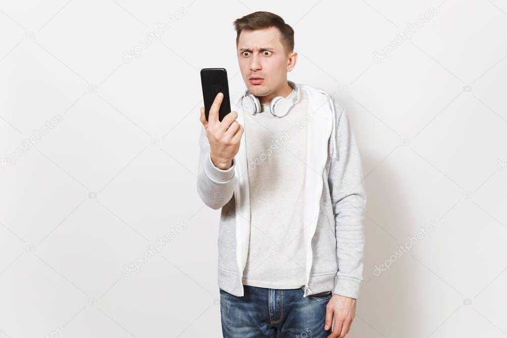 Young sad handsome man student in t-shirt and light sweatshirt with headphones around neck takes his frustrated face on mobile phone isolated on white background. Concept of communication, emotions.