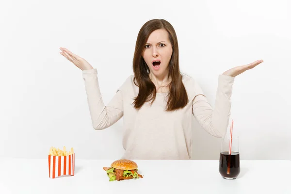 Geïrriteerde vrouw verspreiding handen aan tafel met Hamburger Patat cola in glazen fles geïsoleerd op een witte achtergrond. Goede voeding of Amerikaanse klassieke fastfood. Gebied met kopie ruimte reclame. — Stockfoto