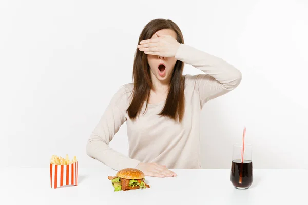 La donna si copre gli occhi con mano a tavola con hamburger, patatine fritte, cola in bottiglia di vetro isolata su fondo bianco. Una corretta alimentazione o un fast food classico americano. Area pubblicitaria con spazio di copia . — Foto Stock