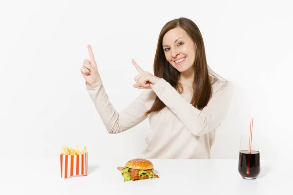 Young woman pointing fingers aside on copy space at table with burger, french fries, cola in glass bottle isolated on white background. Proper nutrition or American classic fast food. Advertising area — Stock Photo, Image