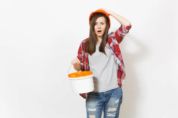 Shocked dissatisfied woman in protective hardhat holding empty paint bucket with copy space and clinging to head isolated on white background. Instrument for renovation apartment. Repair home concept. — Stock Photo, Image
