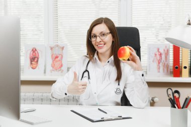 Young smiling female doctor sitting at desk, working on computer with medical documents in light office in hospital. Woman in medical gown with apple in consulting room. Healthcare, medicine concept. clipart