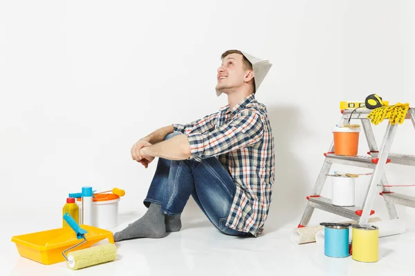 Joven en sombrero de periódico sentado en el suelo con instrumentos para la renovación habitación de apartamentos aislado sobre fondo blanco. Fondo de pantalla, pegar accesorios, herramientas de pintura. Reparar concepto de casa. Vista lateral . — Foto de Stock