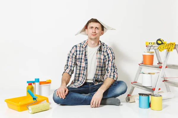 Homem de chapéu de jornal com lápis atrás da orelha sentado no chão com instrumentos para renovação apartamento isolado no fundo branco. Papel de parede, acessórios de colagem, ferramentas de pintura. Conceito de reparação . — Fotografia de Stock