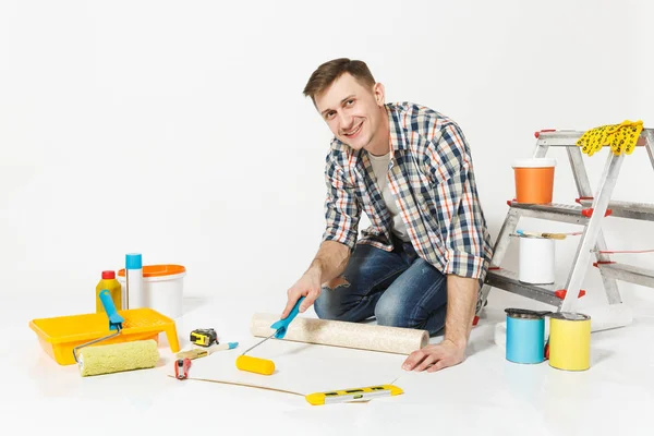 stock image Young man sitting on floor with wallpaper roll, paint roller, instruments for renovation apartment room isolated on white background. Wallpaper, gluing accessories, painting tools. Repair home concept