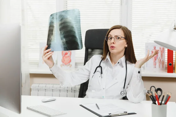 Female doctor sitting at desk, working on computer, holding X-ray of lungs, fluorography, roentgen in light office in hospital. Woman in medical gown, stethoscope in consulting room. Medicine concept.