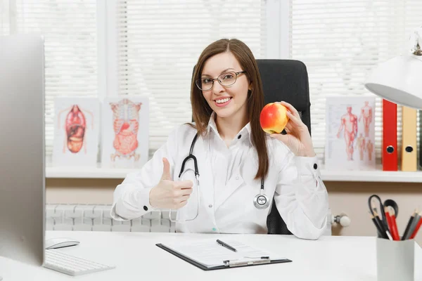 Unga leende kvinnliga läkare sitter vid skrivbord, arbetar på dator med medicinska handlingar i ljus kontor i sjukhuset. Kvinna i medicinsk klänning med apple i samråd rum. Hälsovård, medicin koncept. — Stockfoto