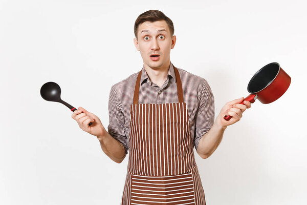 Young surprised man chef or waiter in striped brown apron, shirt holding red empty stewpan black ladle isolated on white background. Male housekeeper or houseworker. Kitchenware and cuisine concept.