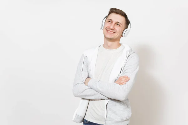 Mladý pohledný usmívající se muž student v tričku a lehká mikina poslouchá hudbu s bílým bezdrátová sluchátka a zbraní přešel hrudníku ve studiu na bílém pozadí. Pojetí emocí — Stock fotografie