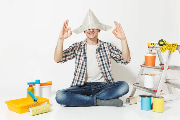 Homem feliz em chapéu de jornal sentado no chão meditando, fazendo ioga, relaxando com instrumentos para renovação apartamento quarto isolado no fundo branco. Papel de parede, ferramentas de colagem. Reparação home concept . — Fotografia de Stock