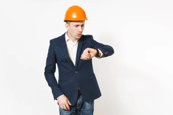 Jovem irritado empresário insatisfeito em terno escuro, capacete laranja construção protetora olhando no relógio na mão isolado no fundo branco. O tempo está a acabar. Trabalhador masculino para publicidade . — Fotografia de Stock