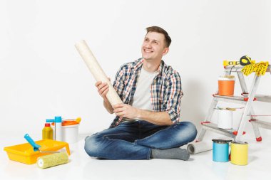 Gülümseyen adam rahat kıyafetler yenileme daire oda beyaz arka plan üzerinde izole için aygıtlar rulo duvar kağıdı ile katta oturuyor. Aksesuarlar, boyama araçlarını yapıştırma. Onarım ev concept.