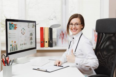 Hastanede ışık ofiste tıbbi belgelerle bilgisayarda çalışan masada oturan genç gülümseyen kadın. Tıbbi önlük, stetoskop Oda danışmanlık içinde kadın doktor. Sağlık, tıp kavramı
