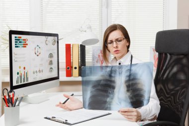 Female doctor sitting at desk, working on computer, holding X-ray of lungs, fluorography, roentgen in light office in hospital. Woman in medical gown, stethoscope in consulting room. Medicine concept. clipart