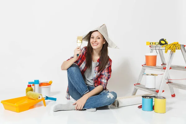 Χαριτωμένο γυναίκα με καπέλο εφημερίδα κάθεται στο πάτωμα με πινέλο, όργανα για ανακαίνιση Διαμέρισμα Δωμάτιο απομονωθεί σε λευκό φόντο. Ταπετσαρία, αξεσουάρ για την κόλληση, εργαλεία ζωγραφικής. Επισκευή concept home. — Φωτογραφία Αρχείου