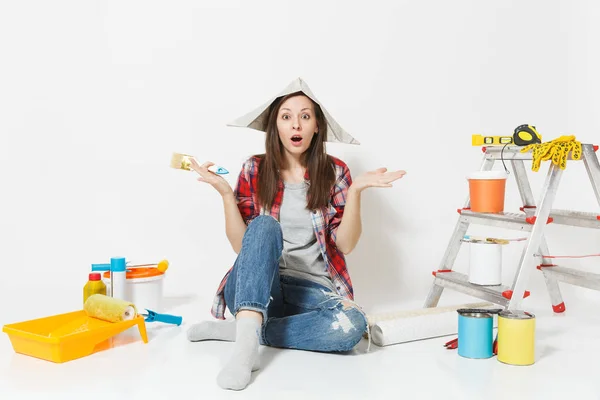 Mulher bonito em chapéu de jornal sentado no chão com escova, instrumentos para renovação apartamento quarto isolado no fundo branco. Papel de parede, acessórios para colar, ferramentas de pintura. Reparação home concept . — Fotografia de Stock