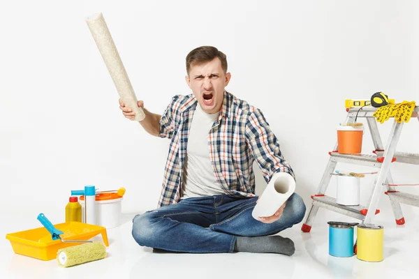 Gritando agresivo joven enojado con rollos de papel pintado, sentado en el suelo con instrumentos para la renovación apartamento aislado sobre fondo blanco. Pegado de accesorios, herramientas. Reparación concepto hogar . — Foto de Stock