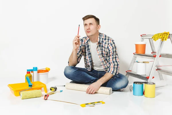 Jovem sentado no chão com papel de parede rolo, lápis, instrumentos para renovação apartamento quarto isolado no fundo branco. Papel de parede, acessórios de colagem, ferramentas de pintura. Reparação home concept . — Fotografia de Stock