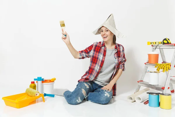 Fun woman in newspaper hat sitting on floor with brush, instruments for renovation apartment room isolated on white background. Wallpaper, accessories for gluing, painting tools. Repair home concept. — Stock Photo, Image