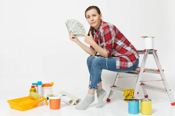 Жінка тримає пакет доларів, готівкові гроші, сидить на сходах з інструментами для ремонту квартири ізольовано на білому тлі. Шпалери аксесуари для склеювання фарбувальних інструментів. Концепція ремонту — стокове фото