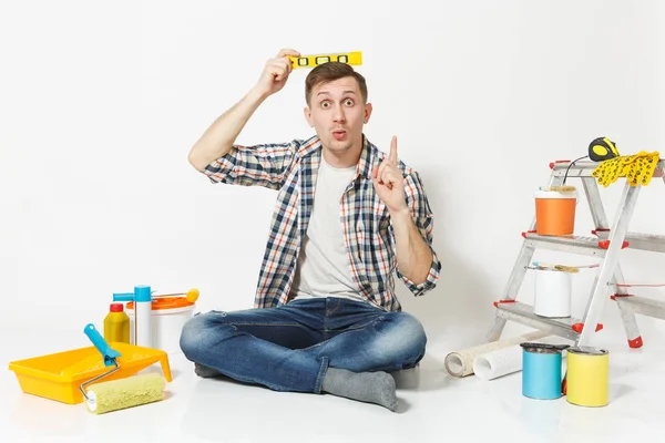 Divertido macho feliz sentado no chão com nível de espírito bolha edifício na cabeça, instrumentos para renovação apartamento isolado no fundo branco. Papel de parede, colando acessórios, ferramentas. Reparação home concept . — Fotografia de Stock