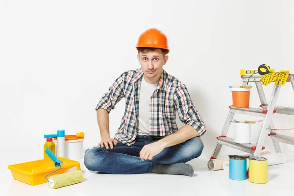 Homem cético em capacete de proteção laranja sentado no chão com instrumentos para renovação apartamento isolado no fundo branco. Papel de parede, acessórios de colagem, ferramentas de pintura. Reparação home concept . — Fotografia de Stock