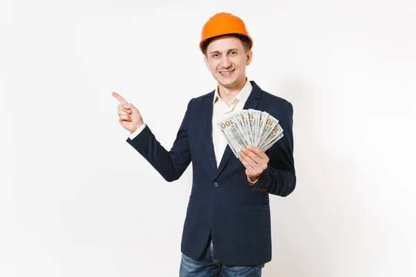 Jeune homme d'affaires souriant en costume sombre, casque de protection tenant paquet de dollars, argent comptant et pointant la main de côté sur l'espace de copie isolé sur fond blanc. Travailleur masculin pour la publicité . — Photo