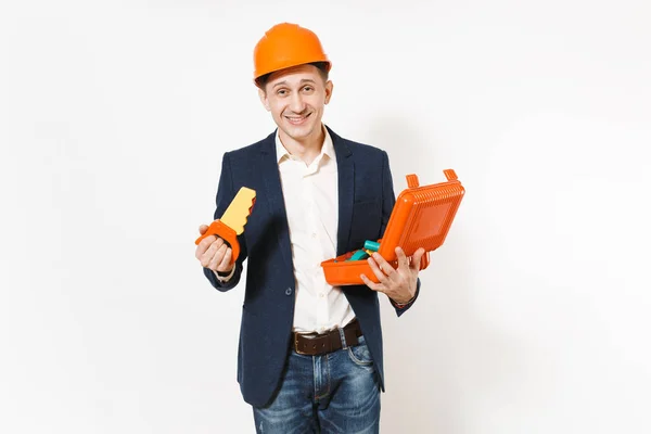 Jovem homem de negócios sorridente em terno escuro, construção protetora capacete laranja segurando caso aberto com instrumentos ou caixa de ferramentas e serra de brinquedo isolado no fundo branco. Trabalhador masculino para publicidade . — Fotografia de Stock