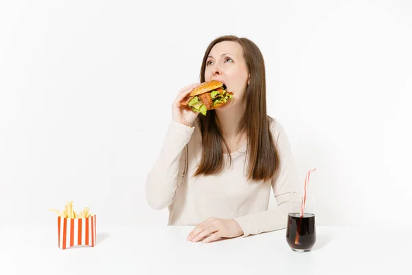 Hungrige junge Frau isst gierig Burger, setzt sich an den Tisch mit Pommes, Cola in Flaschen isoliert auf weißem Hintergrund. richtige Ernährung oder klassisches amerikanisches Fast Food. Werbefläche mit Kopierfläche. — Stockfoto