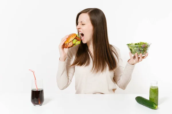 Vrouw met groene detox smoothies, salade met glazen kom, komkommer, eet gretig hamburger, cola in fles geïsoleerd op een witte achtergrond. Goede voeding, gezonde levensstijl, fastfood, dieet concept. — Stockfoto