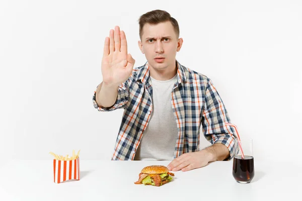 Hombre severo mostrando gesto de alto con palma, sentado a la mesa con hamburguesa, papas fritas, cola en vidrio aislado sobre fondo blanco. Nutrición adecuada o comida rápida clásica americana. Área con espacio de copia . — Foto de Stock