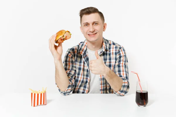 Tânărul frumos arată degetul mare în sus, stă la masă cu burger, cartofi prăjiți, cola în sticlă izolată pe fundal alb. Nutriție adecvată sau fast-food clasic american. Spațiu publicitar cu spațiu de copiere — Fotografie, imagine de stoc