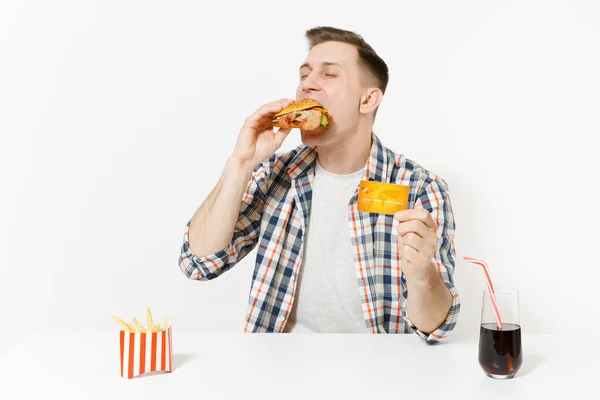 Bonito jovem sentado à mesa com cartão de crédito, hambúrguer, batatas fritas, cola em vidro isolado no fundo branco. Nutrição adequada ou fast food clássico americano. Área de publicidade com espaço de cópia — Fotografia de Stock
