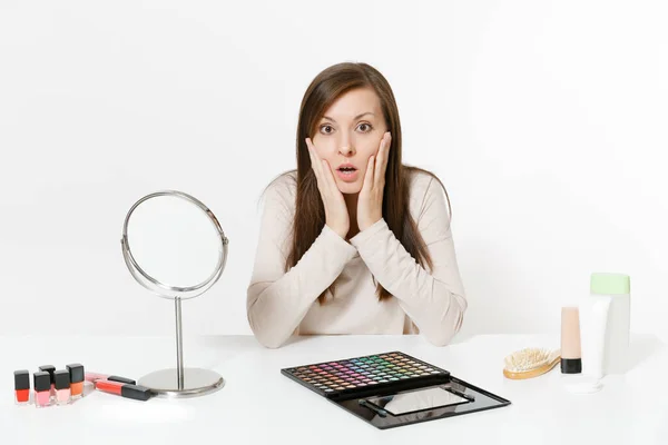 Sorprendida joven molesta sentada a la mesa aplicando maquillaje con cosméticos decorativos faciales establecidos aislados sobre fondo blanco. Belleza concepto de estilo de vida femenino. Área de publicidad con espacio de copia . — Foto de Stock